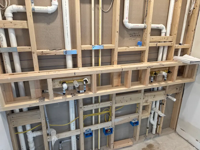 Open bathroom wall framed for a double vanity, with new drain and supply lines and two brass wall-mount faucet valves roughed in