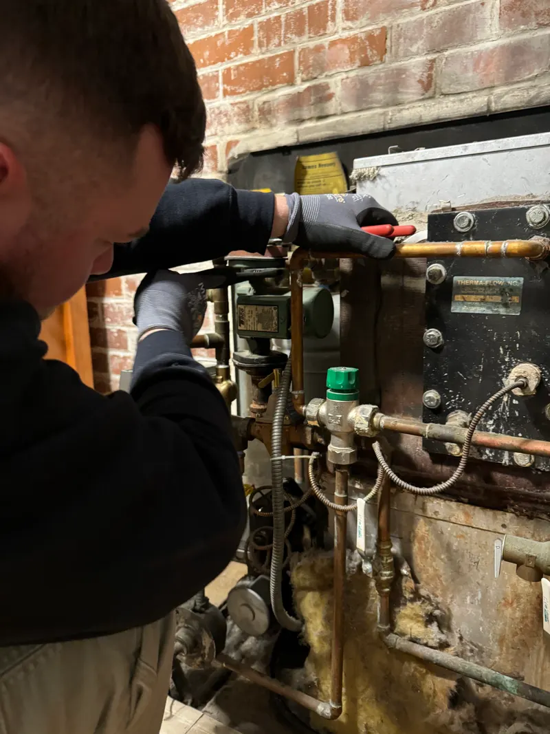 Brandon working on the controls and piping of an older boiler in a basement with a brick wall