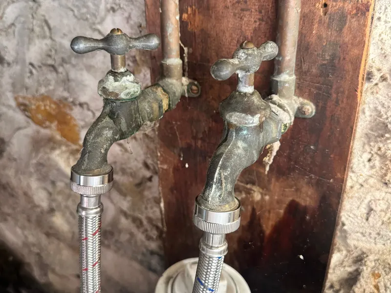 Two corroded old hose bibbs feeding the washing machine hoses, on a board against a stone foundation wall