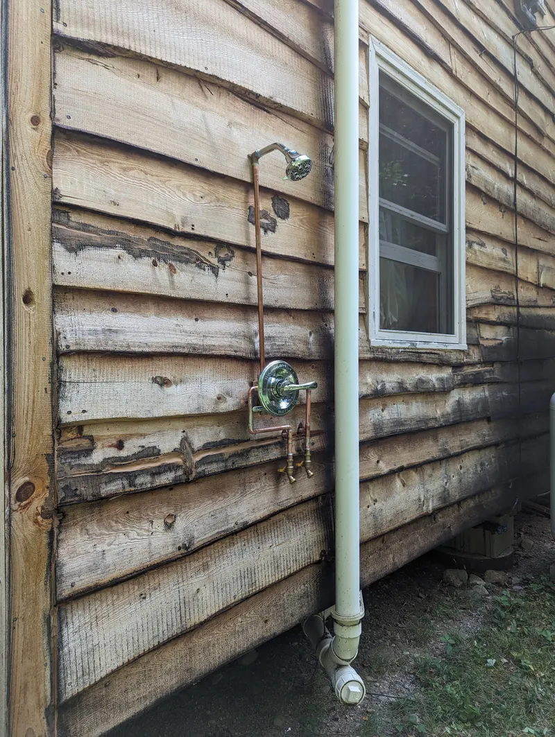 Outdoor shower plumbed on weathered wood siding, with exposed copper supply lines, a shower valve and a shower head