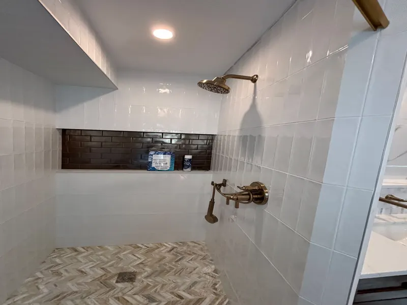 Finished walk-in shower with white tile, a long dark tile niche, a herringbone mosaic floor, and a brushed-brass shower head and handheld shower