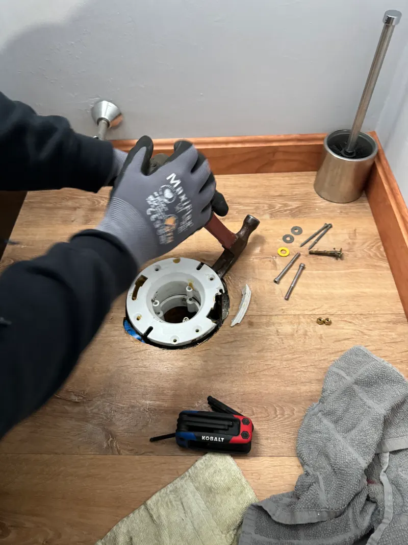 Gloved hands fitting a new toilet flange in the floor, with bolts and tools laid out on a drop cloth