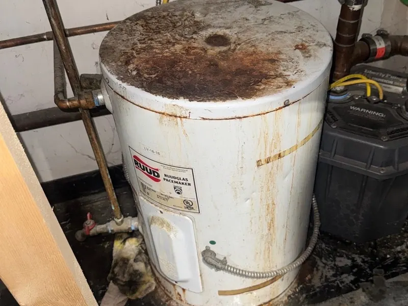 Old Ruud electric water heater with a rust-caked top and rust streaks down the side, in a tight utility corner